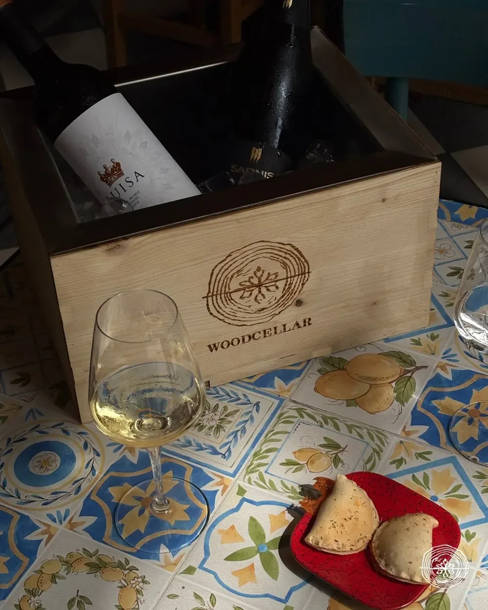 Glacetta Wood Cellar con due bottiglie di vino su un tavolo di maioliche, accanto a un calice di vino bianco.