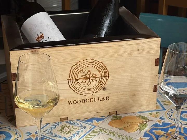Glacetta Wood Cellar con vino bianco e spumante su un tavolo di maioliche, tra due calici di vino bianco.